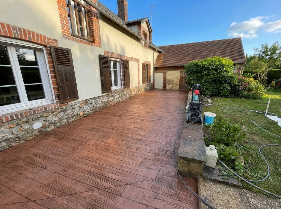 Terrasse extérieure récemment réalisée en béton imprimé façon bois, coloris brun acajou, longeant une maison normande en brique et silex