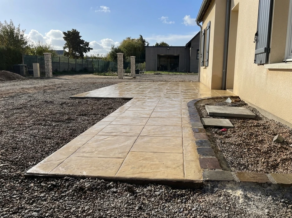 Terrasse extérieure en cours de finition, béton imprimé teinté beige sable, joints quadrillés réguliers, bordure arrondie en pavés multicolores type vieilli